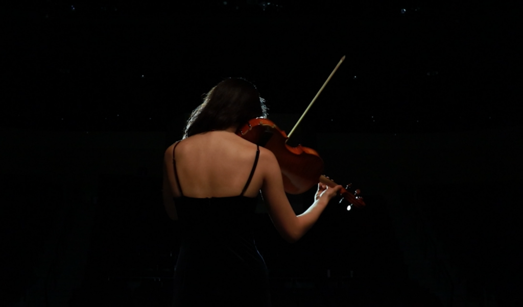 Film still from student film "I Am Blue" featuring a violinist playing from behind and a dark background